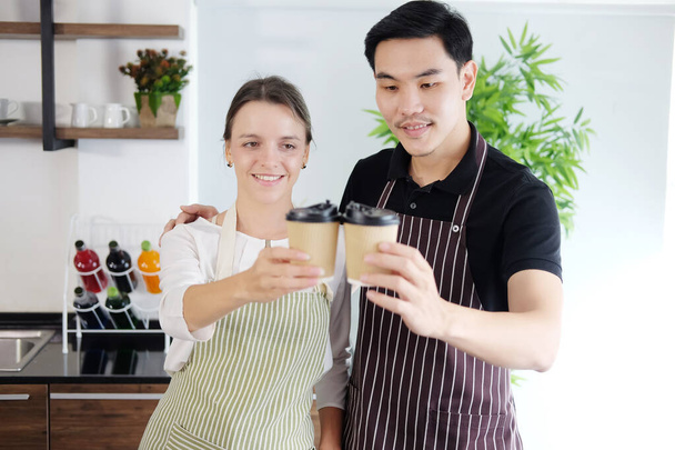 Uśmiechnięta młoda para baristów kocha Azjatę i białą kobietę trzymających kubki gorącej kawy razem. Uruchomienie kawiarni i koncepcji biznesu kawiarni. - Zdjęcie, obraz