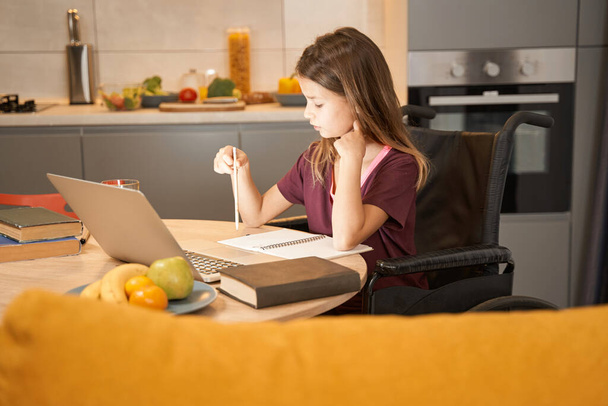 Girl in wheelchair studying with a laptop at kitchen table - Photo, image