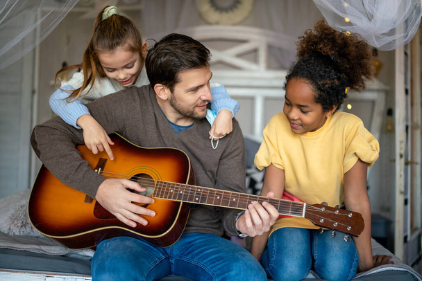 Smiling father with multiethnic children having fun and playing guitar at home. Family love concept - 写真・画像