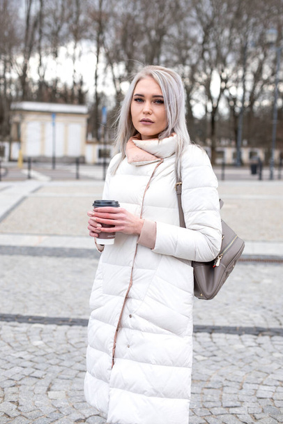 Vertical blonde attractive woman, staying look at camera, in warm white jacket, leather bag pack, drinking cup of coffee - Photo, Image