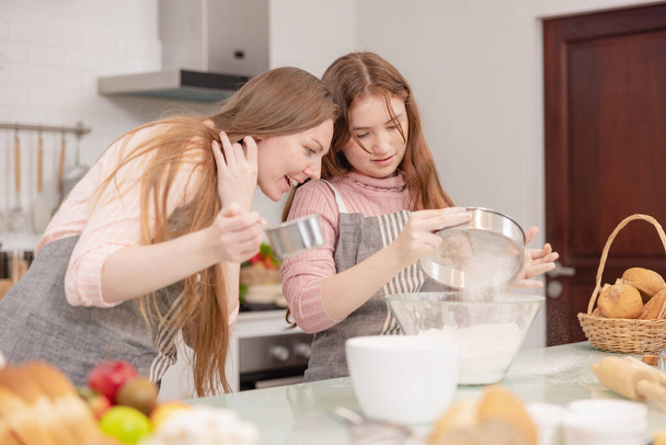 Mom and her younger kids daughter in a modern kitchen area celebrating joyous learning, baking, cake decoration, happy small daughter make prepare sweet pastries with caring mom on weekend at home - Photo, Image