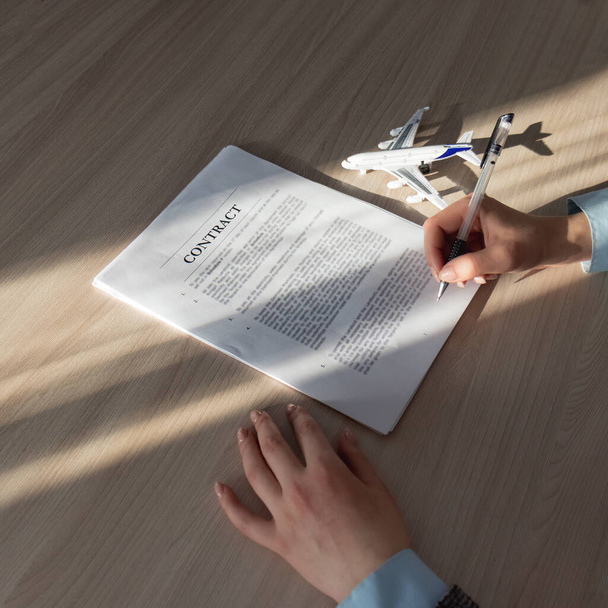 Flight insurance, travel insurance concept. contract aircraft. aircraft and air equipment leasing. Close-up. Women's hands hold a pen and study the contract before signing, next to the document lies the symbol of the aircraft and the sunbeam shines.  - Фото, зображення