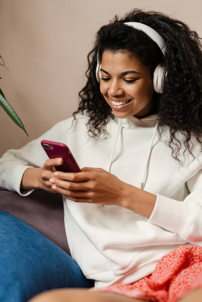 Young black woman in headphones using cellphone while sitting on sofa at home - Foto, afbeelding