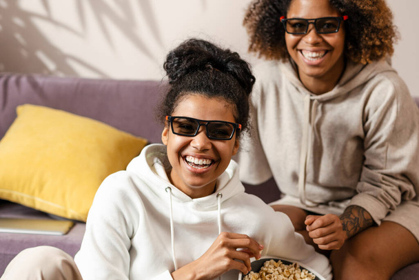 Two happy multiethnic women friends sitting on a couch at home with snacks watching TV laughing - Fotoğraf, Görsel