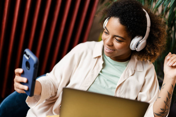 Nuori musta nainen kuulokkeissa kannettavan tietokoneen ja matkapuhelimen avulla kotona - Valokuva, kuva