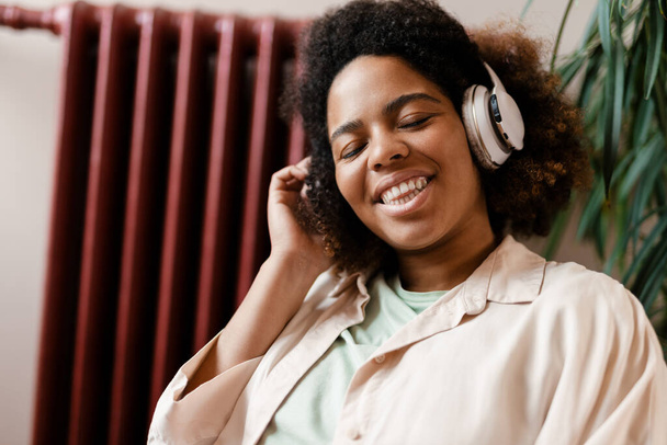 Young black woman smiling while listening music with headphone at home - Фото, изображение