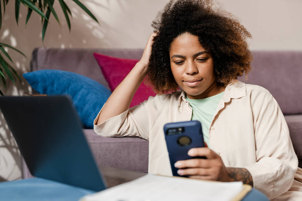 Young black woman using mobile phone while working with laptop at home - Photo, image