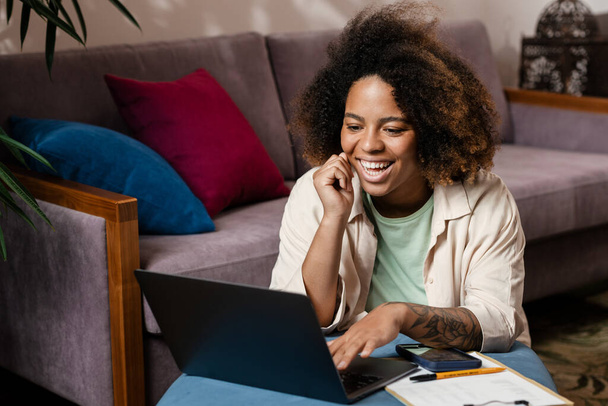Young black woman smiling and using laptop while sitting on floor at home - Foto, Bild
