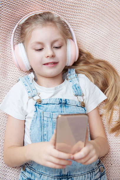 Маленька дівчинка з мобільним телефоном слухає музику, лежачи в ліжку
 - Фото, зображення