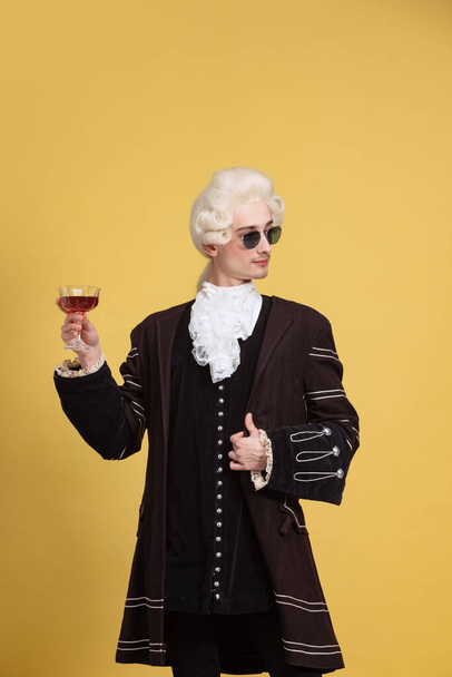 Portrait of young elegant man in white wig and vintage medieval outfit posing isolated on yellow background. Art, beauty, fashion - Foto, Imagen