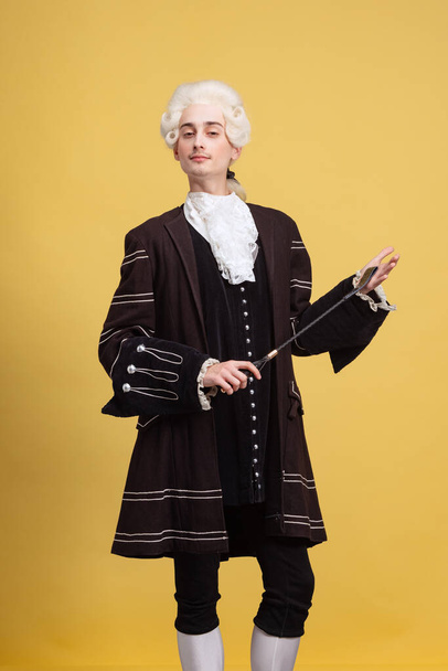 Portrait of young elegant man in white wig and vintage medieval outfit posing isolated on yellow background. Art, beauty, fashion - 写真・画像