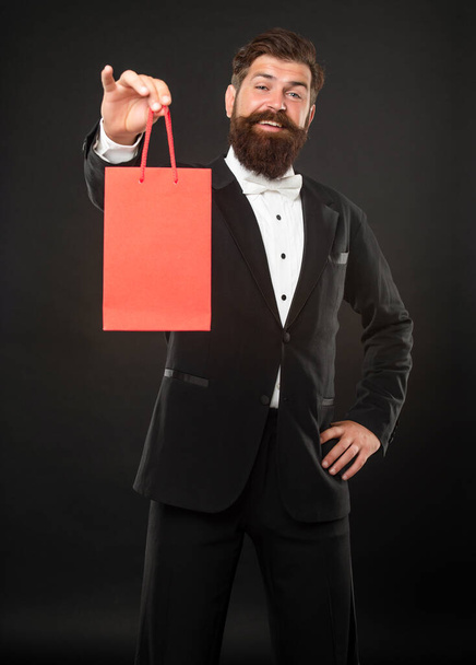 happy man in tuxedo bow tie on black background presenting shopping bag for mens day. copy space. - Foto, afbeelding