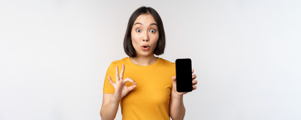 Eccitato ragazza asiatica che mostra lo schermo del telefono cellulare, segno ok, raccomandare app per smartphone, in piedi in maglietta gialla su sfondo bianco - Foto, immagini