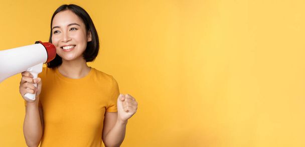 Sorrindo asiático mulher de pé com megafone, anunciando smth, publicidade produto, de pé sobre fundo amarelo - Foto, Imagem