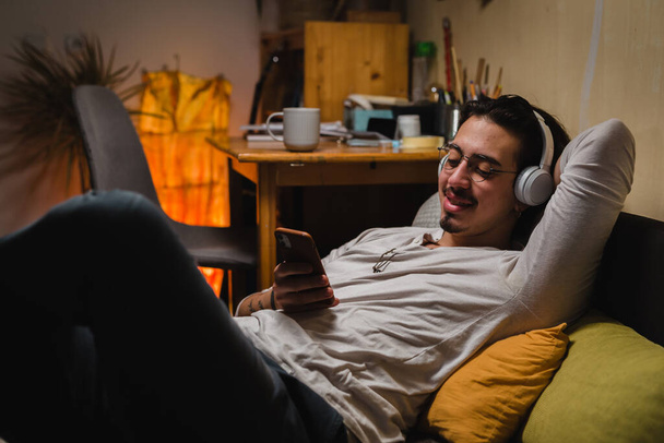 Man relaxing on sofa at home playing with his phone and wearing headphones - 写真・画像