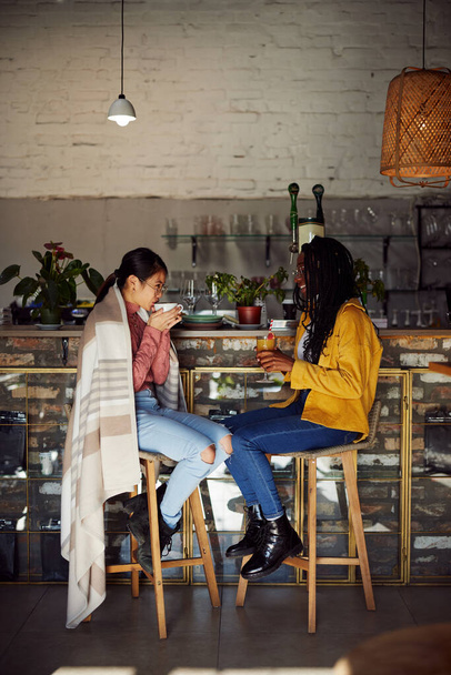 Two multicultural friends sit in the cafeteria, drink beverages, and chat. - 写真・画像