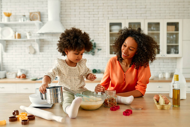 Afroamerikanerin mit ihrer kleinen Tochter mit lockigem, flauschigem Haar, die Spaß hat und in der Küche Gebäck backt. Mutter und Tochter kochen zusammen. - Foto, Bild