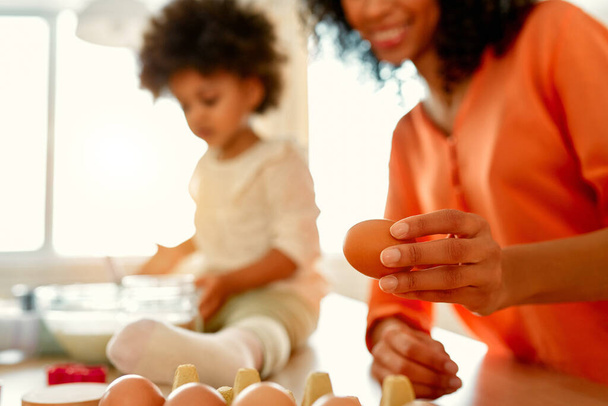 Афроамериканська жінка зі своєю маленькою донькою з кучерявим пухнастим волоссям розважається і готує тістечка на кухні. Мама і дочка готують разом. Жінка дістає яйця з лотка.. - Фото, зображення