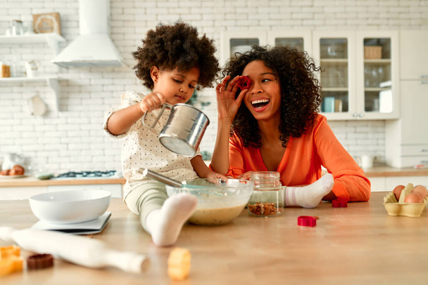 Afro-Amerikaanse vrouw met haar dochtertje met krullend pluizig haar die plezier heeft en gebak kookt in de keuken. Mam en dochter koken samen. Kind zit aan tafel koekjes te bakken. - Foto, afbeelding