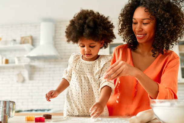 Afro-Amerikaanse vrouw met haar dochtertje met krullend pluizig haar die plezier heeft en gebak kookt in de keuken. Mam en dochter koken samen. Koekjes uitsnijden met koekjesschaar. - Foto, afbeelding