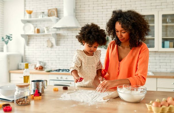 Αφροαμερικανή γυναίκα με την κορούλα της με σγουρά αφράτα μαλλιά που διασκεδάζουν και μαγειρεύουν γλυκά στην κουζίνα. Η μαμά και η κόρη μαγειρεύουν μαζί. Παιδί κόβει μπισκότα με κόφτες μπισκότων. - Φωτογραφία, εικόνα