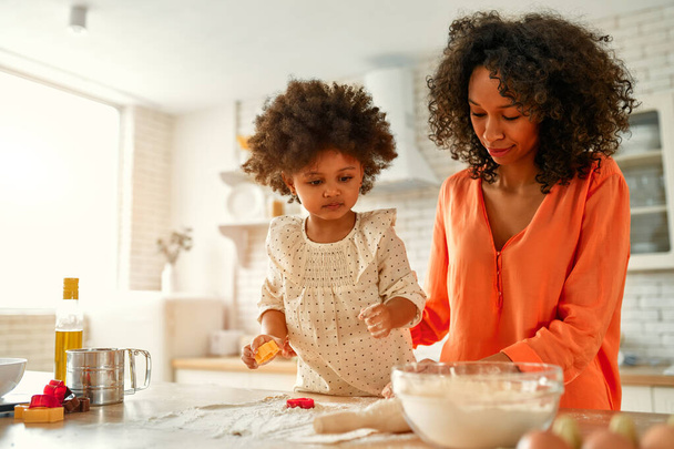 Afro-Amerikaanse vrouw met haar dochtertje met krullend pluizig haar die plezier heeft en gebak kookt in de keuken. Mam en dochter koken samen. Koekjes uitsnijden met koekjesschaar. - Foto, afbeelding
