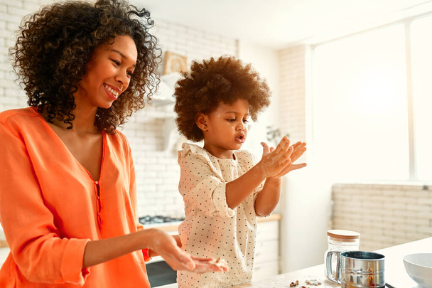 Donna afroamericana con la sua figlioletta dai capelli ricci soffici divertendosi e cucinando dolci in cucina. Mamma e figlia cucinano insieme. Bambino ritaglio biscotti con tagliabiscotti. - Foto, immagini