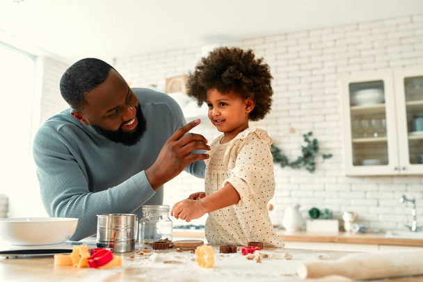 Kıvırcık saçlı ve mutfakta hamur işi pişiren küçük kızları olan Afro-Amerikan bir aile. Baba ve kızı birlikte yemek pişiriyor ve hamurdan kurabiye kesiyorlar.. - Fotoğraf, Görsel