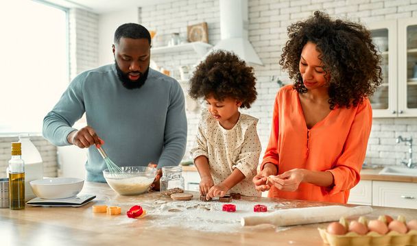Eine afroamerikanische Familie mit ihrer kleinen Tochter mit lockigem Flauschhaar hat Spaß und kocht in der Küche Gebäck. Papa, Mutter und Tochter kochen gemeinsam und schneiden Plätzchen aus dem Teig. - Foto, Bild