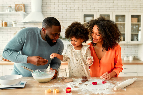 Eine afroamerikanische Familie mit ihrer kleinen Tochter mit lockigem Flauschhaar hat Spaß und kocht in der Küche Gebäck. Papa, Mutter und Tochter kochen gemeinsam und schneiden Plätzchen aus dem Teig. - Foto, Bild