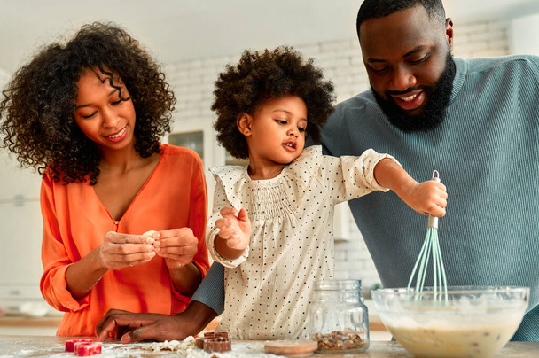 Afroamerykańska rodzina z córeczką z kręconymi puszystymi włosami bawiącymi się i gotującymi wypieki w kuchni. Tata, mama i córka robią razem ciasteczka.. - Zdjęcie, obraz