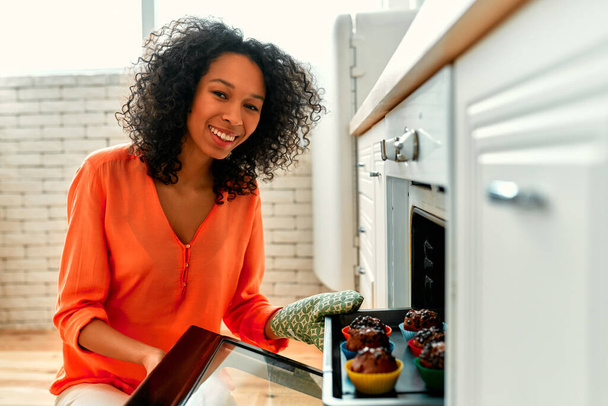 African American cute woman with curly fluffy hair taking out a baking sheet with baked muffins from the oven. - Valokuva, kuva
