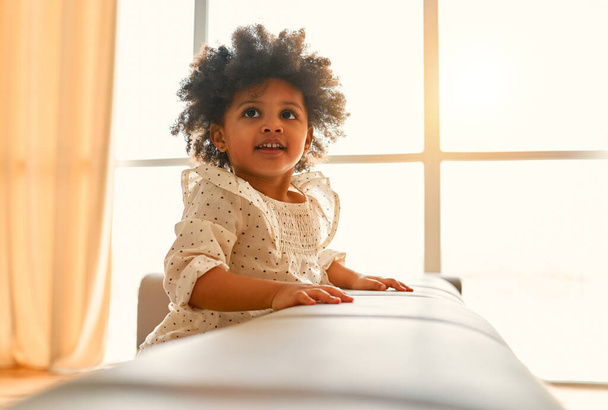 Aranyos afro-amerikai lány göndör bolyhos haj ül a kanapén egy ruhát a nappaliban otthon. - Fotó, kép
