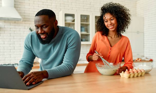 Linda pareja afroamericana preparar pasteles dulces para el desayuno y pasar tiempo romántico juntos en la cocina en casa utilizando el ordenador portátil. - Foto, imagen