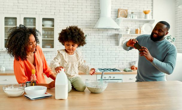 Mulher afro-americana com sua filhinha com cabelo encaracolado fofo se divertindo e cozinhando na cozinha. Mãe e filha cozinham juntas enquanto o pai tira uma foto em um smartphone. - Foto, Imagem