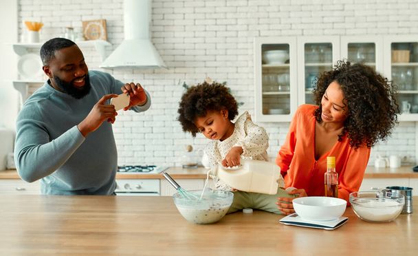 Mujer afroamericana con su hija pequeña con el pelo rizado esponjoso divertirse y hornear en la cocina. Mamá e hija cocinan juntas mientras papá toma una foto en un teléfono inteligente. - Foto, Imagen