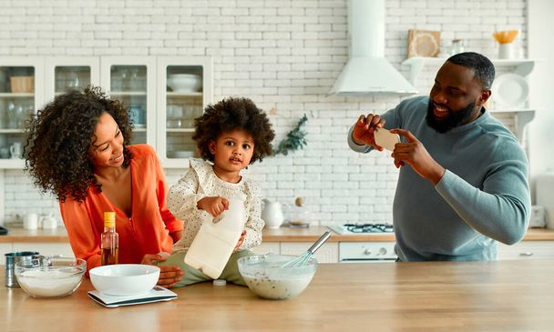 Femme afro-américaine avec sa petite fille aux cheveux bouclés et duveteux qui s'amuse et cuit dans la cuisine. Maman et fille cuisinent ensemble pendant que papa prend une photo sur un smartphone. - Photo, image