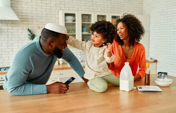 Афро-американская семья со своей маленькой дочерью с вьющимися пушистыми волосами, веселящимися и пекущимися на кухне. Маленькая девочка положила тарелку на голову своего отца и смеется. - Фото, изображение