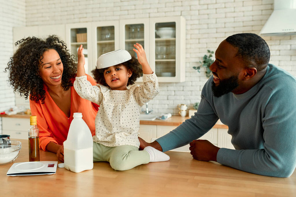 Eine afroamerikanische Familie mit ihrer kleinen Tochter mit lockigem Flauschhaar hat Spaß beim Backen in der Küche. Das kleine Mädchen legt sich den Teller auf den Kopf und lacht. - Foto, Bild