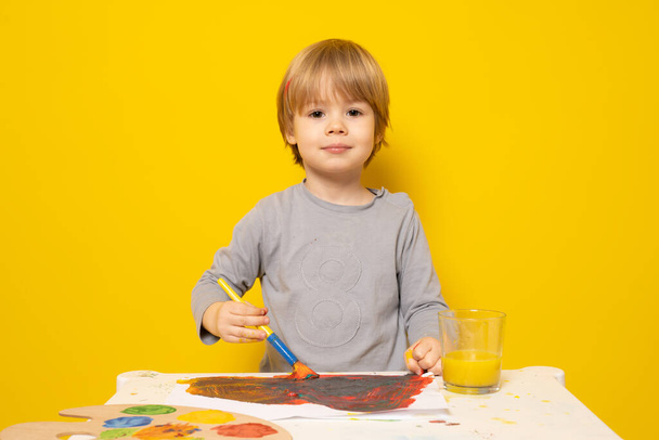 Όμορφο αγοράκι κάθεται στο γραφείο και ζωγραφίζει πολύχρωμη εικόνα με μπογιά και πινέλα πάνω από κίτρινο φόντο. Η έννοια των παιδιών. - Φωτογραφία, εικόνα