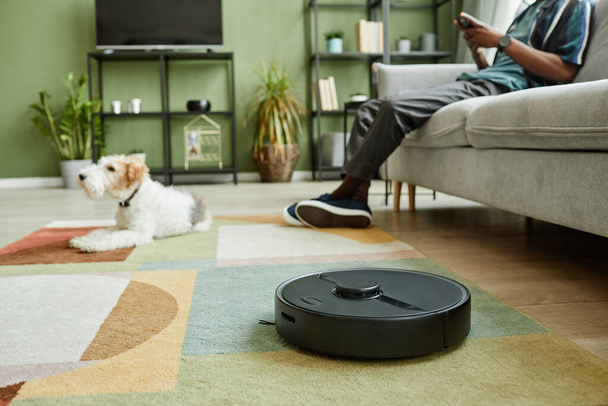 Close up of robot vacuum cleaner on carpet with young man relaxing in background, copy space - Photo, Image