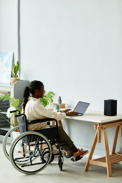 Comprimento total mínimo da jovem mulher negra em cadeira de rodas usando laptop enquanto trabalhava no escritório em casa, espaço de cópia - Foto, Imagem