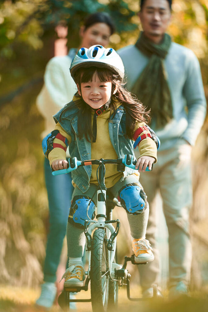 kleines asiatisches Mädchen mit Helm und Vollschutzausrüstung radelt im Stadtpark, während die Eltern von hinten zusehen - Foto, Bild