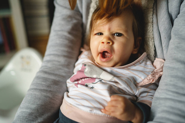 Μικρό καυκάσιο μωρό στα χέρια της μητέρας του να κάθεται και να κοιτάζει την κάμερα να ουρλιάζει νυσταγμένη - Φωτογραφία, εικόνα