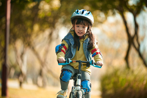 piękny szczęśliwy Azji mała dziewczynka z kask i pełna ochrona bieg jazda na rowerze w parku miejskim - Zdjęcie, obraz