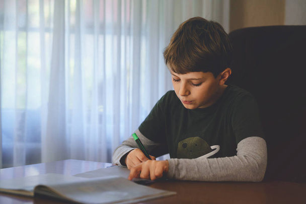 Hard-working happy school kid boy making homework during quarantine time from corona pandemic disease. Healthy child writing with pen, staying at home. Homeschooling concept - Photo, Image