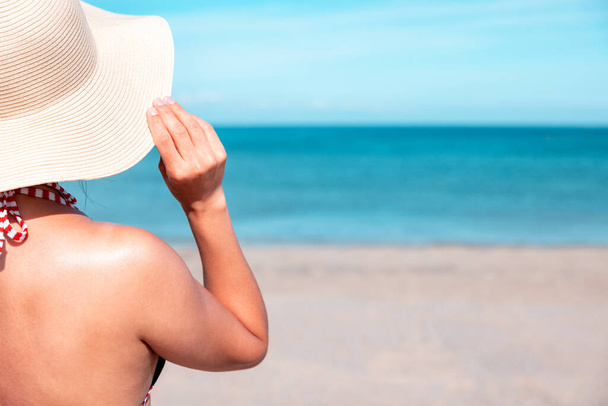 Woman at seaside in Cornwall looking at sea and holding straw hat - Rear view of a woman standing on the beach with ocean on background on a sunny day - Travel and happiness concepts - Foto, imagen