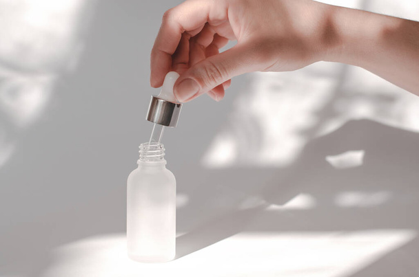 Female hand holding pipette with essential oil or serum. Natural cosmetics for body care. White cosmetic bottle and female hand on a white background with shadows - Photo, Image