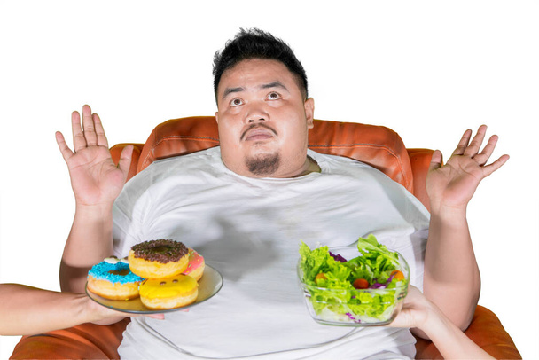 Obesity man expressing confused while choosing a vegetable salad or donuts in the studio. Isolated on white background - Valokuva, kuva