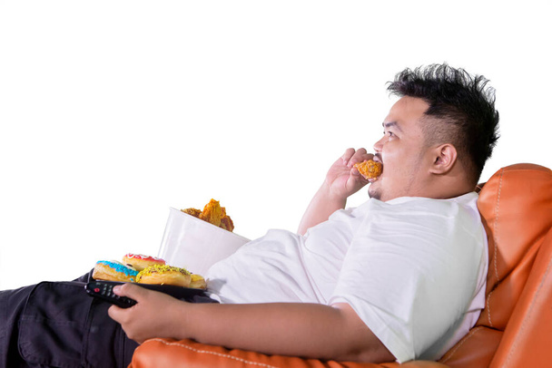 Человек с ожирением ест пончики и жареную курицу на кресле во время просмотра телевизора в студии. Изолированный на белом фоне - Фото, изображение